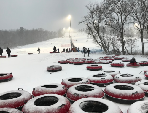 Snow-covered tubing hill with people climbing and sliding down, surrounded by red inflatable tubes and trees.