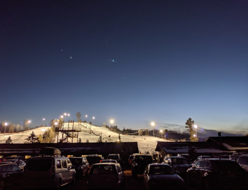 A snowy hill with ski lights at dusk, surrounded by parked cars and a clear blue sky.