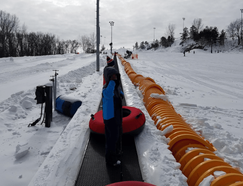 A snowy hill with children waiting in line to go tubing on a conveyor belt, surrounded by snow-covered trees.