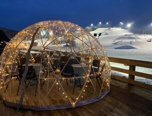 A cozy, illuminated igloo on a snowy ski slope, with chairs inside and lights twinkling against a night sky.