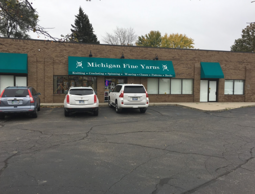 Exterior of Michigan Fine Yarns store with green awnings, parked cars, and a clear sky in the background.