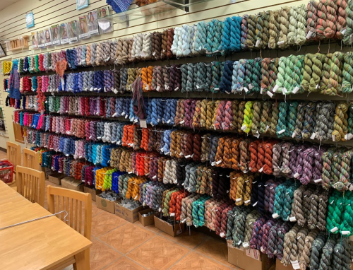 A colorful display of yarn skeins arranged on shelves in a craft store, with wooden tables in the foreground.
