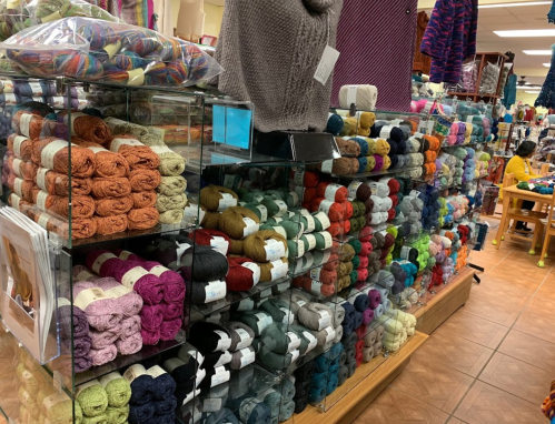 A colorful display of neatly arranged yarn skeins in a craft store, with various textures and shades visible.