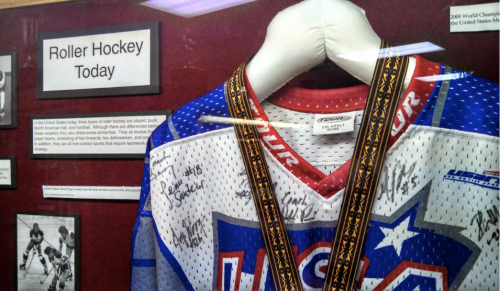 A signed roller hockey jersey displayed in a case, featuring a medal and a sign about roller hockey's history.