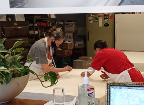 Two women in aprons work together at a table, focused on a task, with plants and a hand sanitizer bottle in the foreground.