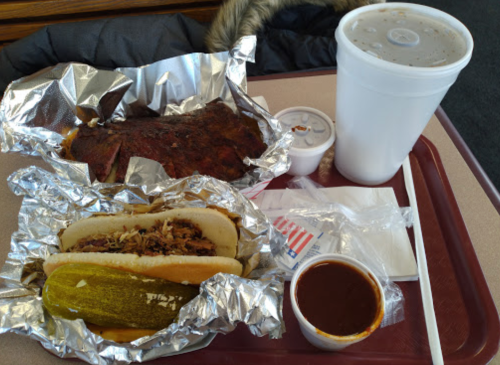 A tray with two foil-wrapped sandwiches, a cup of soda, and a small container of sauce, alongside pickles.