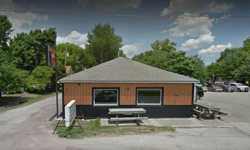 A small, orange and black building with a sloped roof, surrounded by trees and a parking area.