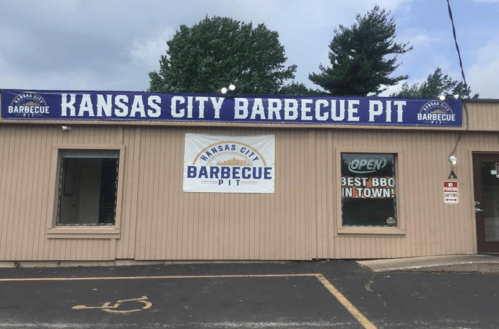 Exterior of Kansas City Barbecue Pit restaurant with a large sign and an "Open" banner. Trees in the background.