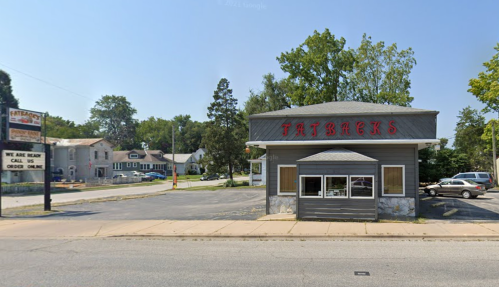 A gray building with "Fatbacks" sign, surrounded by trees and houses, on a sunny day. A sign advertises takeout.