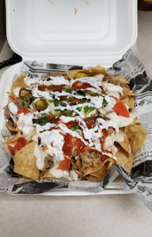 A takeout container filled with nachos topped with pulled pork, sour cream, salsa, jalapeños, and diced tomatoes.