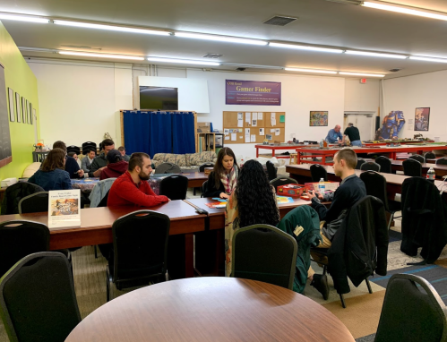 A group of people playing games at tables in a spacious community room with colorful walls and bulletin boards.