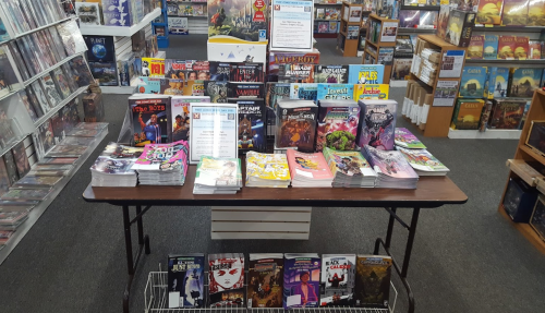 A display table in a store featuring various comic books and board games, with shelves of games in the background.