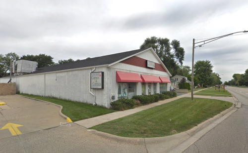 A white building with red accents and awnings, located on a grassy corner lot beside a road.