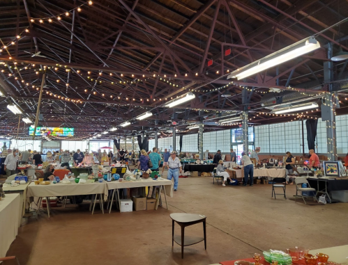A bustling indoor market with tables displaying various items, people browsing, and string lights overhead.