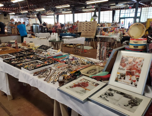 A bustling market scene with tables displaying various vintage jewelry, collectibles, and framed sports memorabilia.
