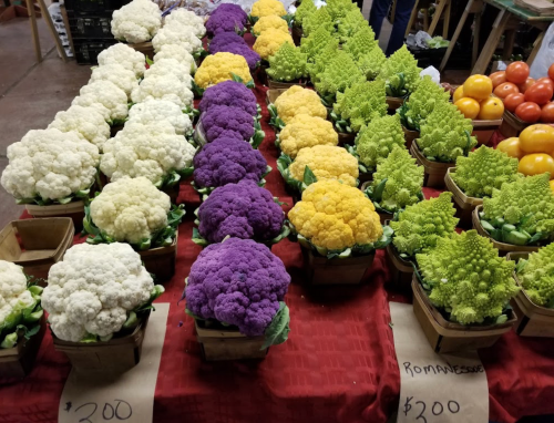 Colorful display of cauliflower and romanesco in wooden baskets at a market, with prices labeled.