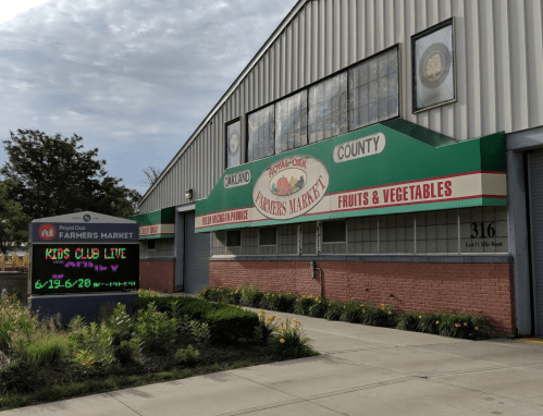 Exterior of a farmers market building with a sign promoting "Kids Club Live" and displaying fruits and vegetables.