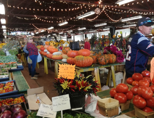 A bustling market with colorful pumpkins, flowers, and fresh produce, decorated with string lights.