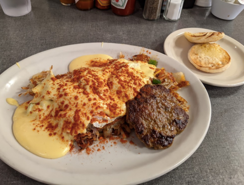 A plate of hash browns topped with eggs, sausage patty, and creamy sauce, served with a biscuit on the side.