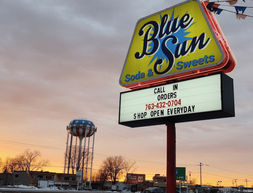 Sign for Blue Sun Soda & Sweets with a colorful sunset and a water tower in the background. Call-in orders displayed.