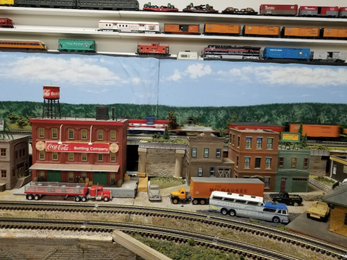 A model train setup featuring buildings, vehicles, and a backdrop of trees, with trains running on tracks above.