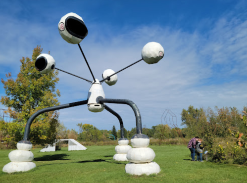 A large, abstract sculpture with multiple spherical elements stands in a park, while two people tend to plants nearby.