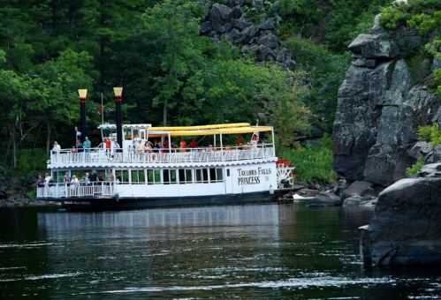 A white paddleboat with passengers floats on a calm river, surrounded by lush green trees and rocky cliffs.
