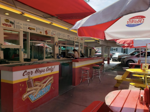 A colorful outdoor diner with red and yellow decor, featuring car hop service and shaded seating areas.