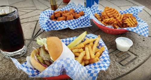 A burger with lettuce and pickles, crinkle fries, and two baskets of fried snacks on a table with a drink.