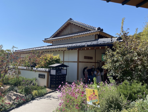 A traditional-style building surrounded by greenery and flowers, with people gathered near the entrance on a sunny day.