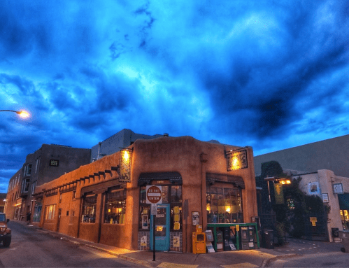 A charming adobe building with large windows under a dramatic blue sky at dusk. Street signs and greenery are visible.