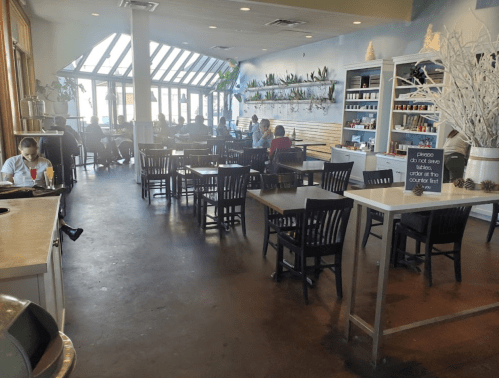 Bright café interior with tables, chairs, and patrons enjoying their meals. Large windows let in natural light.