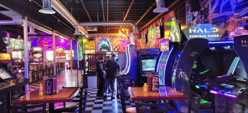 A vibrant arcade with colorful lights, featuring gaming machines and tables, and two people interacting near the games.