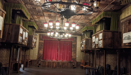 Interior of a vintage theater with wooden decor, green curtains, and a red stage curtain, illuminated by hanging lights.