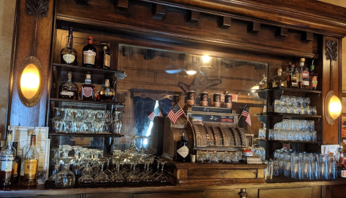 A wooden bar with shelves of liquor bottles, glasses, and a vintage cash register, reflecting a warm, inviting atmosphere.