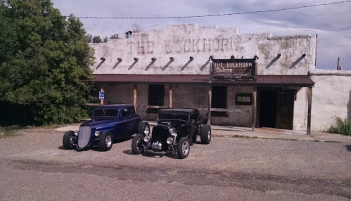 Two vintage cars parked in front of an old building with a sign reading "The Buckhorn Saloon." Green trees in the background.