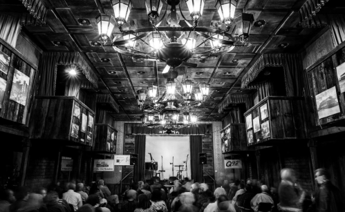 A dimly lit venue with a chandelier, featuring a stage set for a performance and an audience in motion.