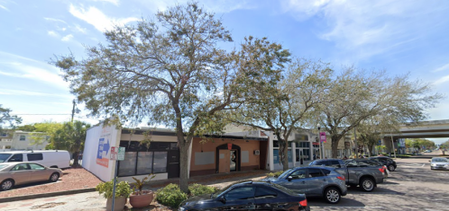 A street view of a commercial building with trees and parked cars under a clear blue sky.