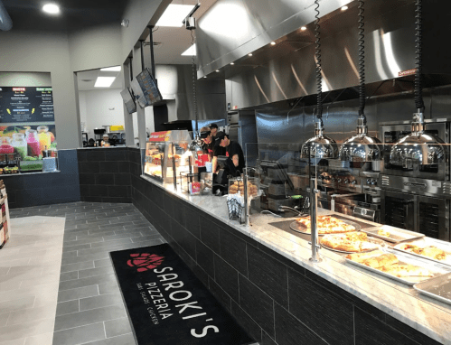 A modern pizzeria interior with a counter, staff preparing food, and a display of pizzas under warm lighting.