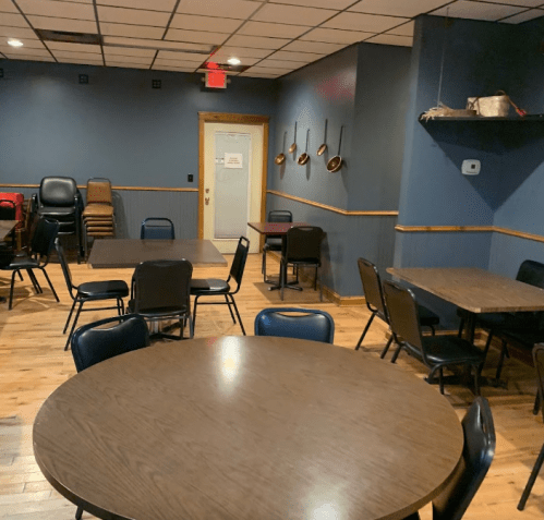 A dimly lit room with several round and square tables, stacked chairs, and a door at the back.