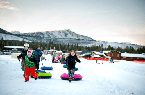 Children and adults joyfully tubing down a snowy hill, with mountains and a winter landscape in the background.