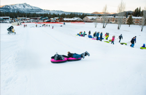 Two people slide down a snowy hill on inflatable tubes, with others waiting in line behind them. Snowy mountains in the background.