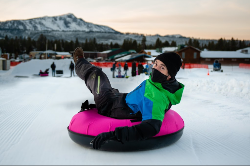 A person in winter gear sits on a pink snow tube, smiling against a snowy landscape and mountains in the background.