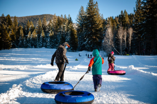 Two children and an adult walk on snow with colorful snow tubes, surrounded by trees and a clear blue sky.