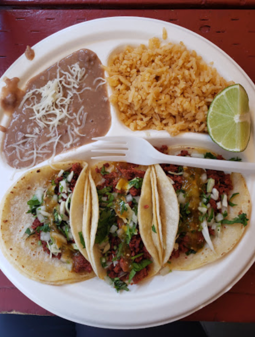 A plate with three tacos topped with onions and cilantro, rice, refried beans, and a lime wedge.
