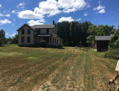 A large two-story house with a porch, surrounded by green trees and a grassy field under a blue sky with clouds.