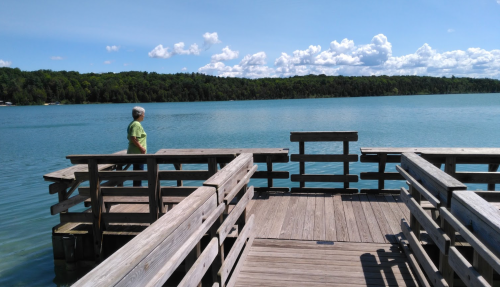 A person stands on a wooden dock overlooking a calm lake under a blue sky with fluffy clouds.