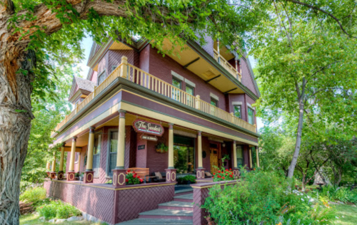 A charming three-story house with a wraparound porch, surrounded by lush greenery and colorful flowers.