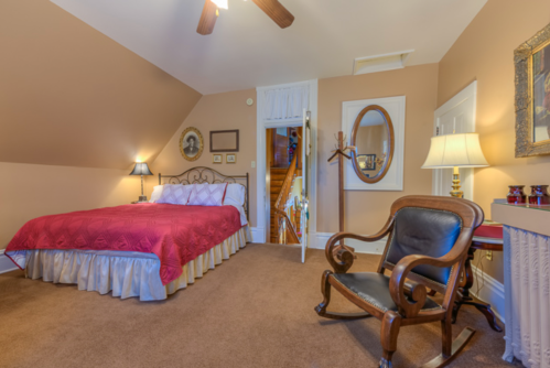 Cozy bedroom with a red bedspread, rocking chair, and warm lighting, featuring a sloped ceiling and wooden accents.