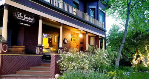 A charming purple house with a porch, surrounded by greenery and flowers, illuminated by soft lights in the evening.